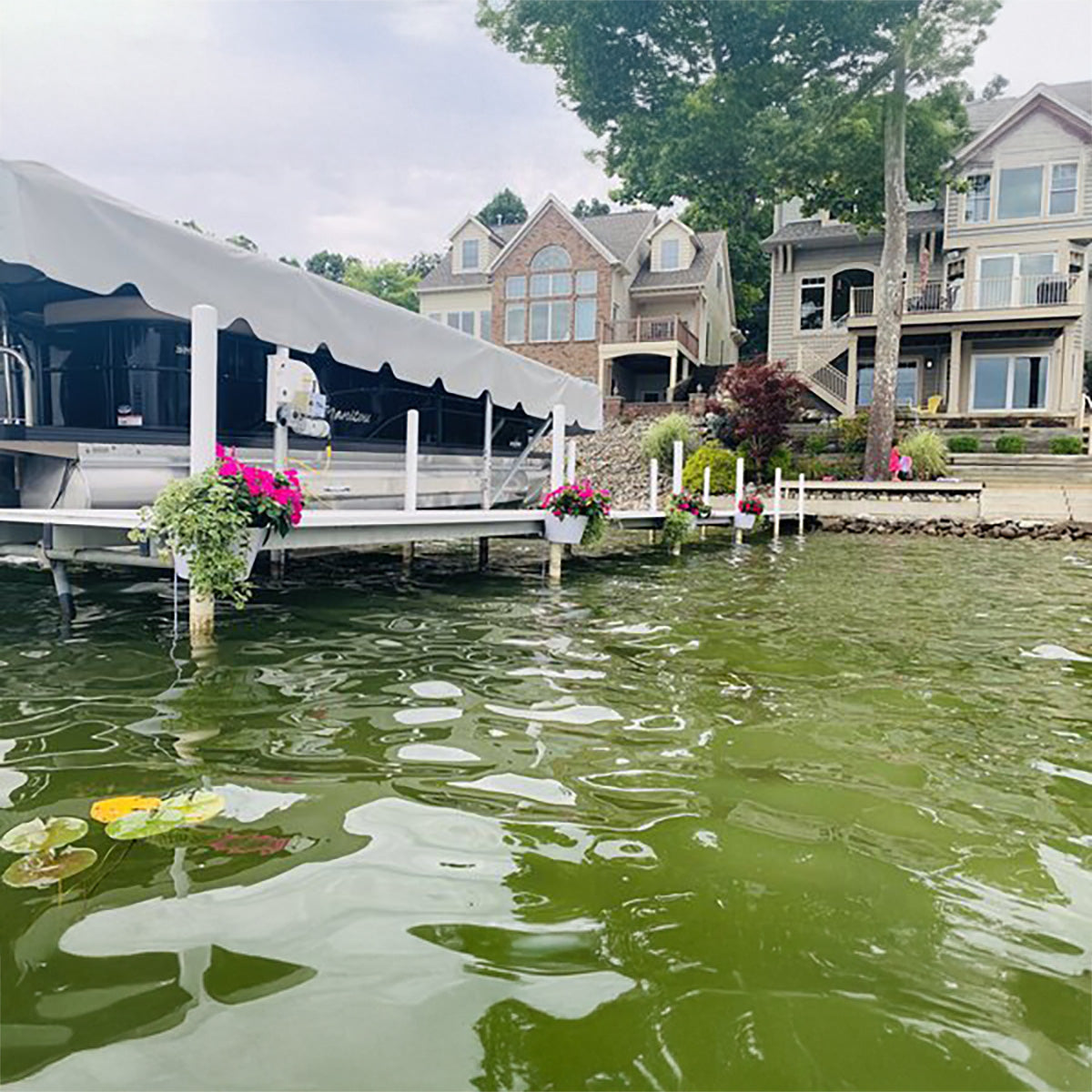 Dock with a covered boat, flowers, and houses in the background on a lake.
