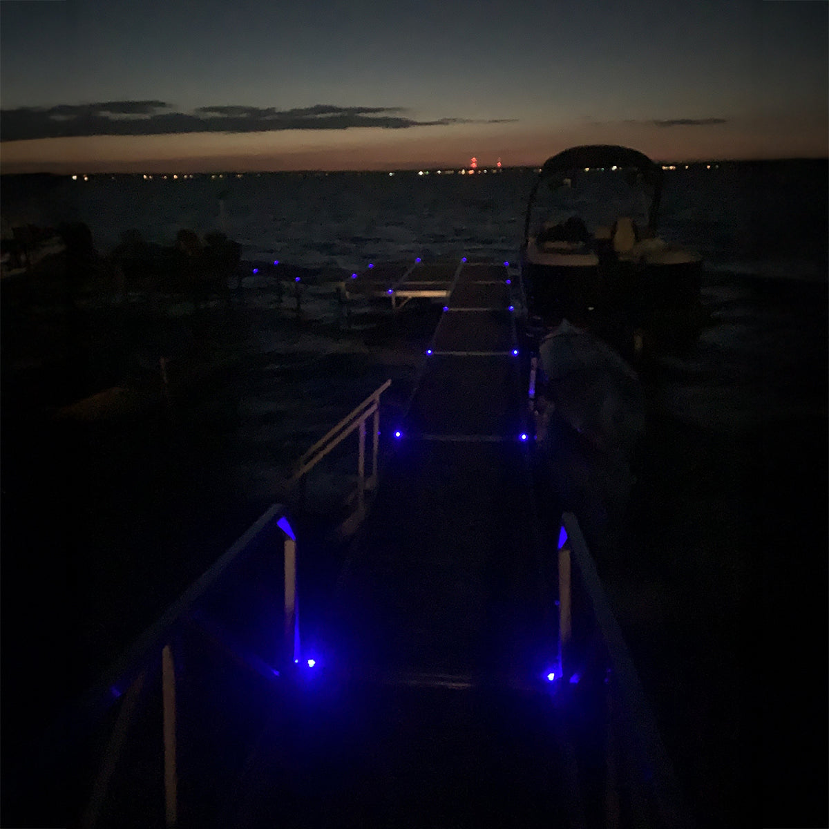 Dock with a boat at night, illuminated by blue lights, with a city skyline in the distance.