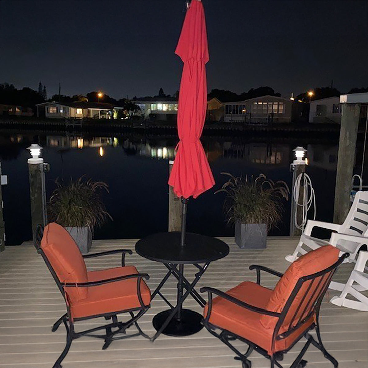 Outdoor patio set with orange chairs, black table, and red umbrella on a dock at night.