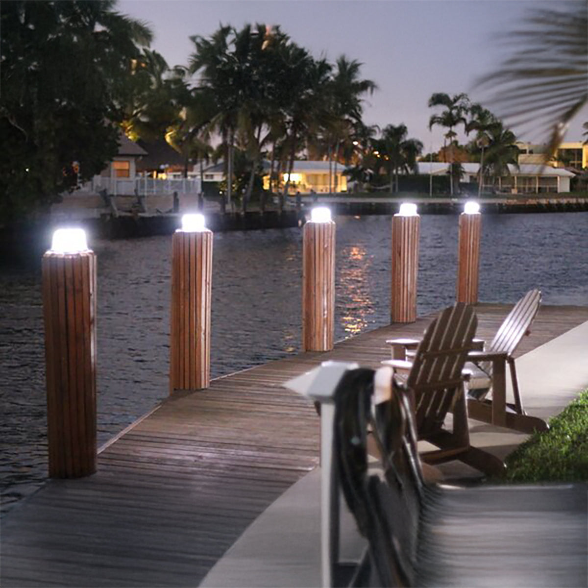 Wooden dock with illuminated posts and lounge chairs by a waterfront at night.