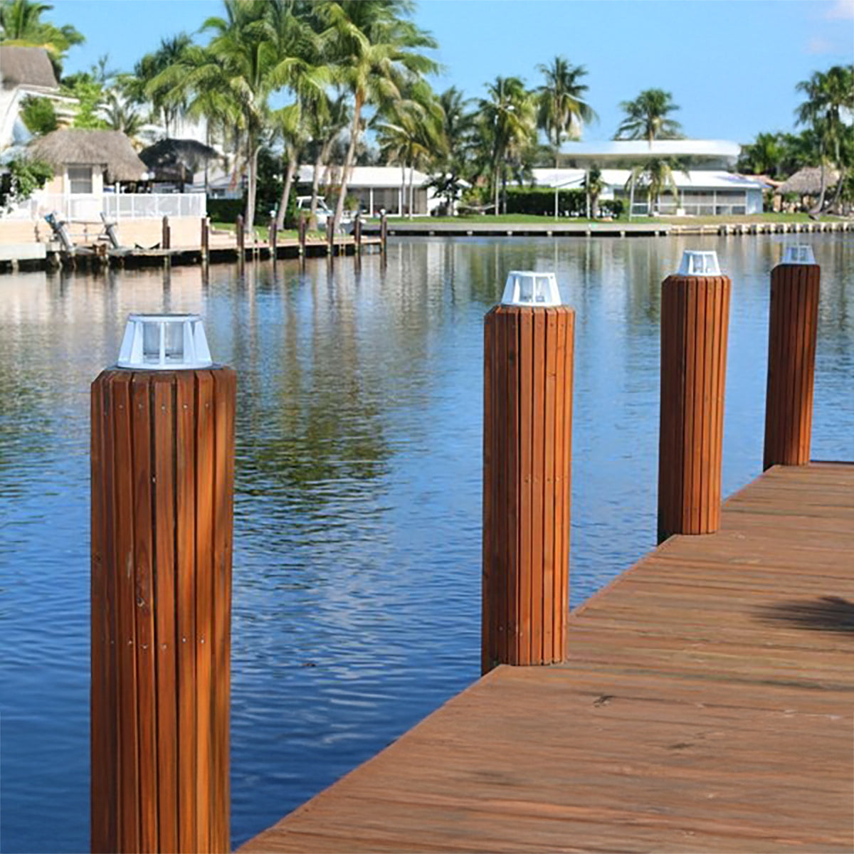 Wooden dock with solar lights on posts by a calm body of water with palm trees in the background.