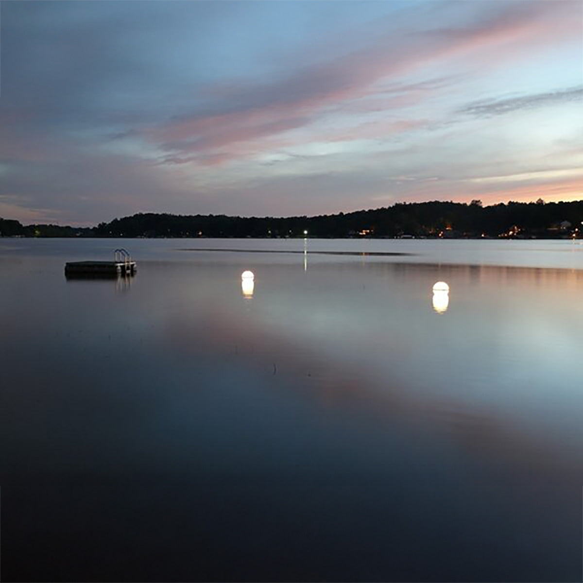 Dusk over a lake with a dock and lights reflecting on the water.