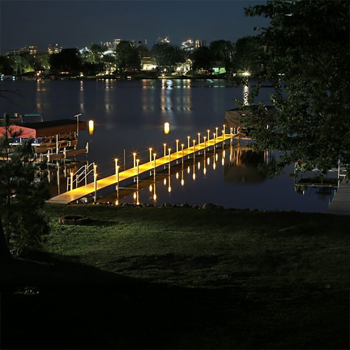 Evening scene of a dock with lights reflecting on water, surrounded by trees.