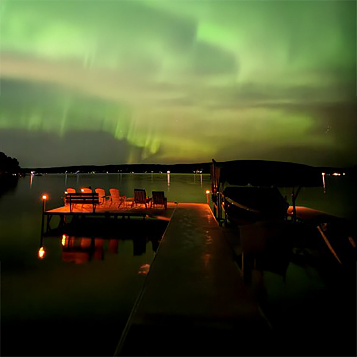 A dock with chairs under a vibrant green aurora borealis sky over a lake.