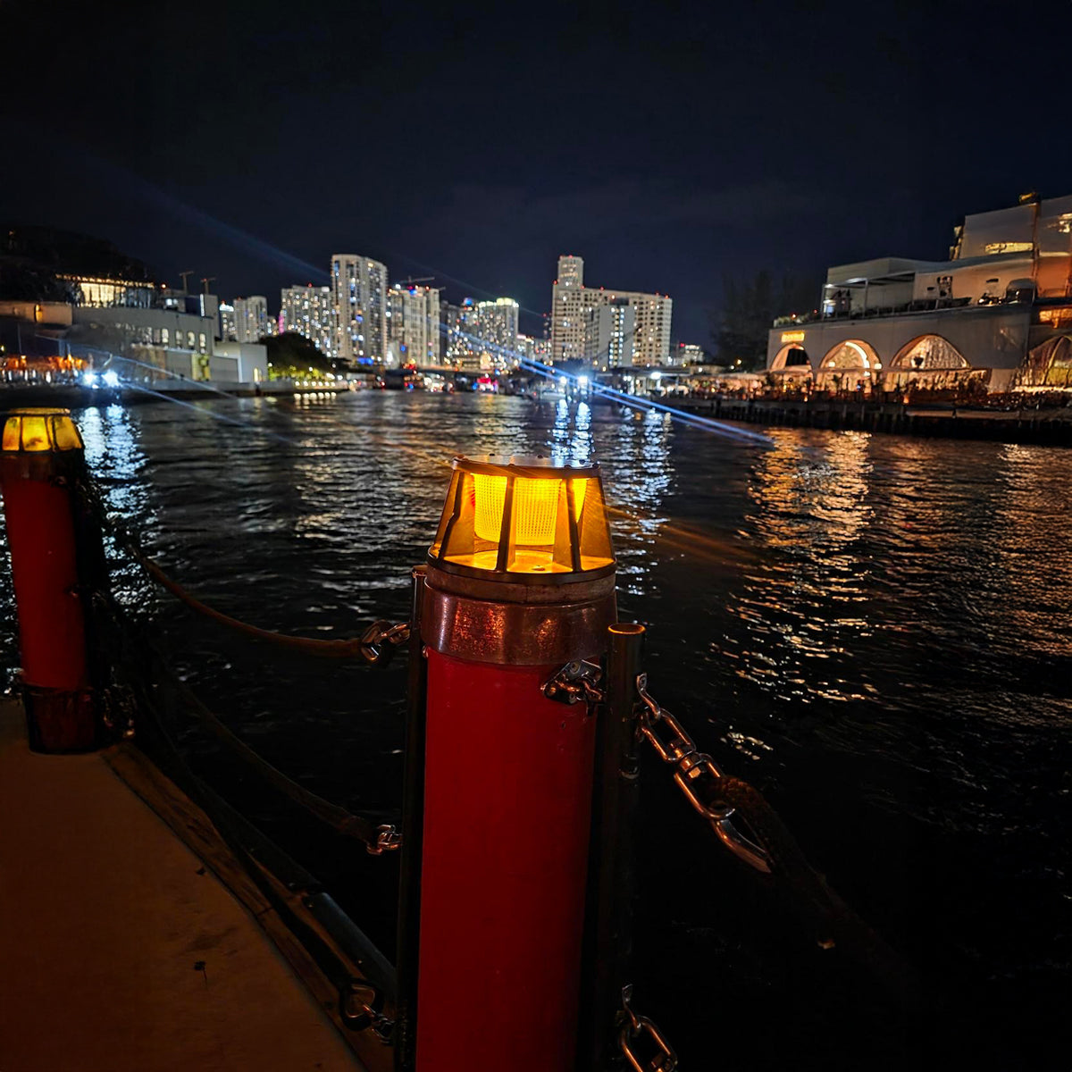 Cityscape at night with illuminated buildings reflected in water, featuring red and yellow light posts.