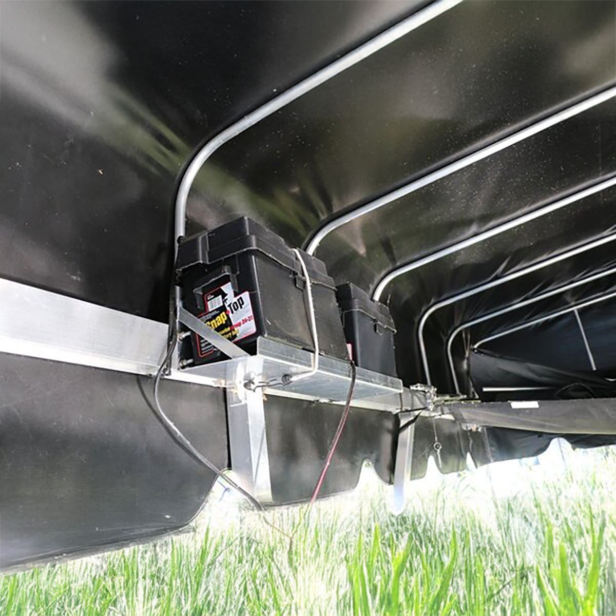 Two batteries mounted on a metal bracket under a vehicle with grass in the background.