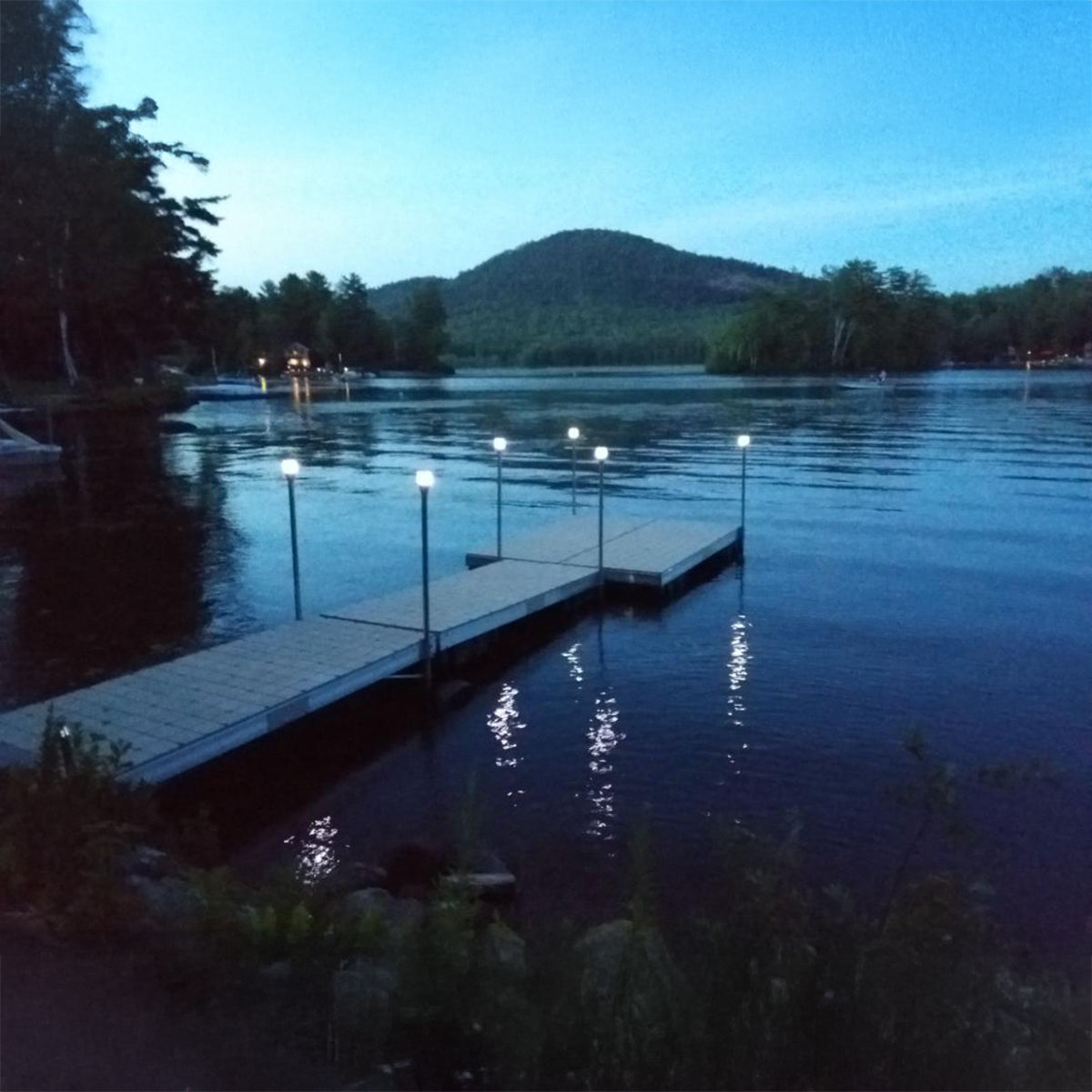 Evening scene at a dock with lights on a lake surrounded by trees and mountains.