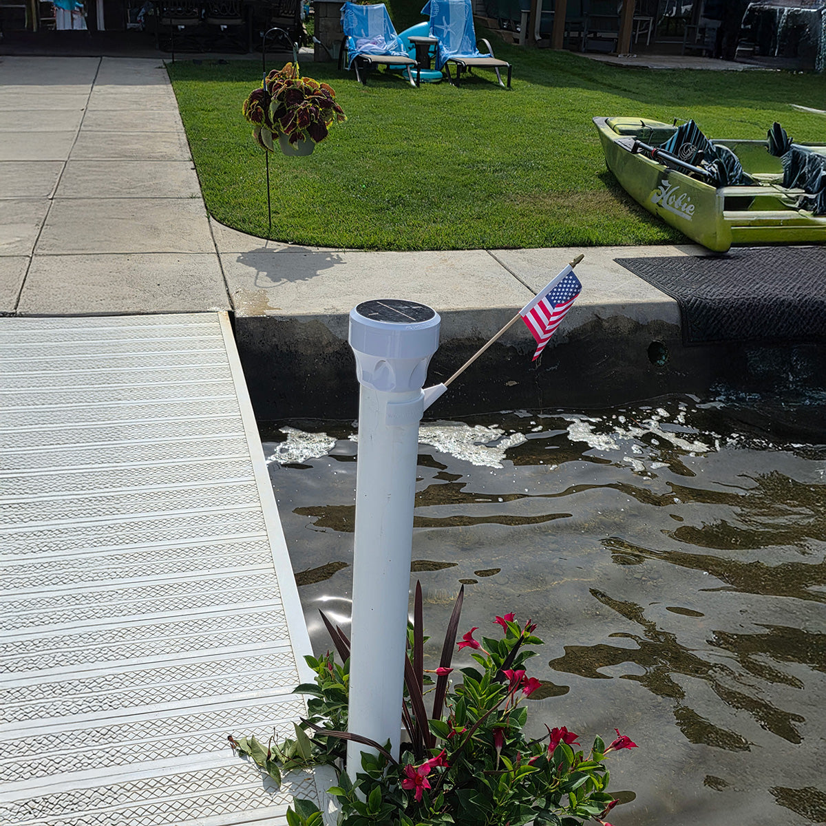 White decorative column with a solar light on top, surrounded by plants and a flag, near a pool area.