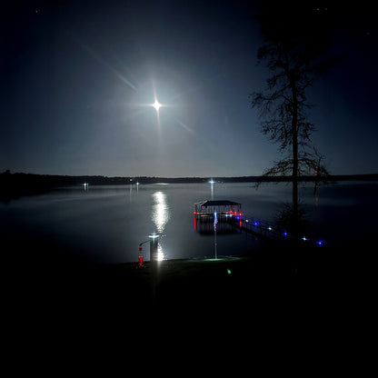 Distant light source over a lake at night with a dock and tree silhouette.