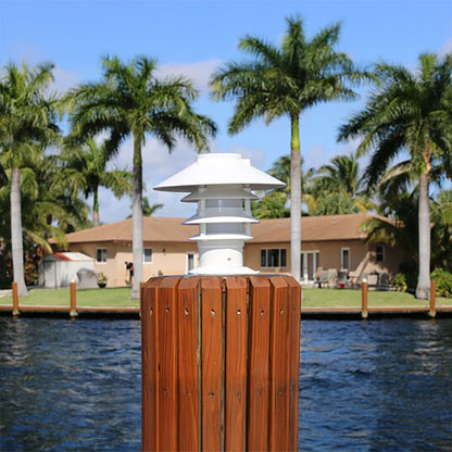 Wooden dock post with a white lamp against a backdrop of palm trees and a house.
