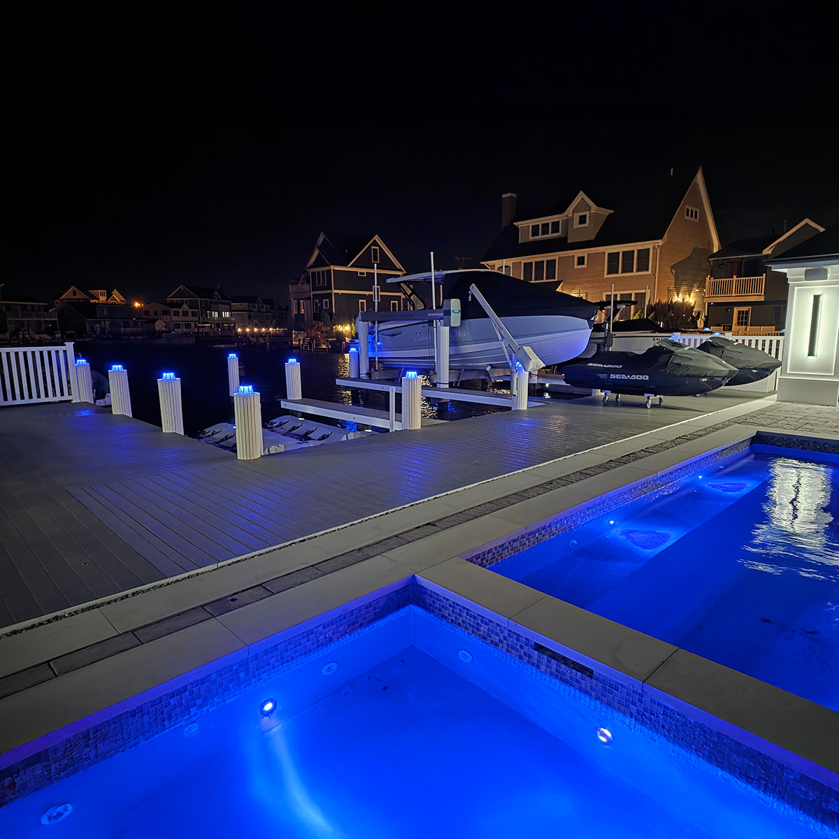 Pool area with illuminated blue lights at night, featuring a dock with boats and houses in the background.